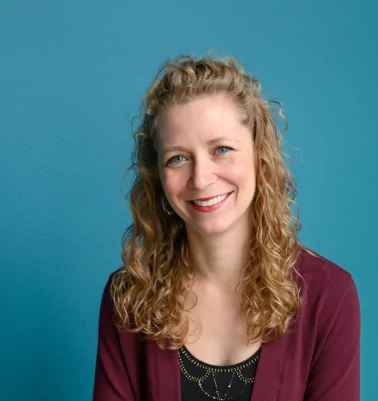 a photo of Johanna smiling with wavy blond hair, blue eyes, and a maroon cardigan in front of a teal wall